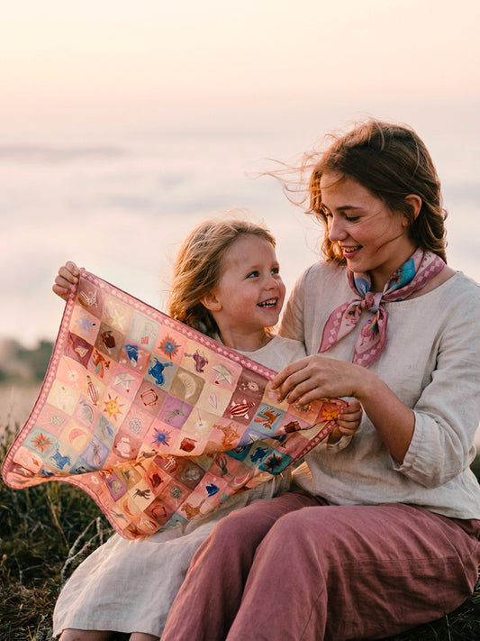 A close-up shot of two young sisters sitting on a grassy hill above a sea of clouds. They are examining the whimsical pastel illustrations on a Journey of Yula scarf. The older girl wears a small, neatly tied 45cm neckerchief. Soft morning light, natural skin textures, and cinematic atmosphere emphasizing a 'pocket of joy' moment.