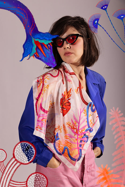 A stylish woman wearing a "Heart Aflame" silk scarf over a blue blazer, paired with cat-eye sunglasses, showcasing a luxury wearable art look.