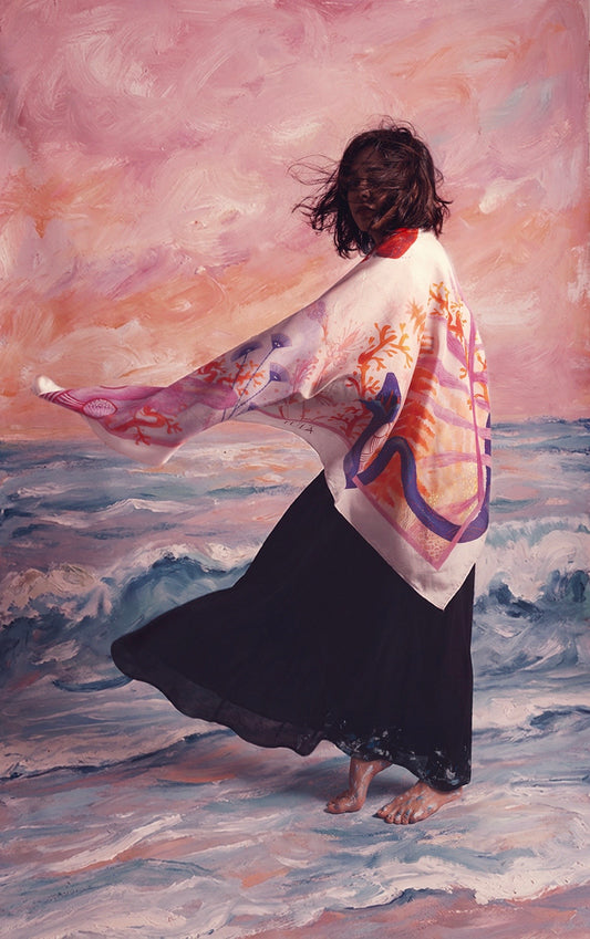 Artistic shot of a large silk scarf billowing in the wind against a painted sunset background, showcasing vibrant coral and botanical illustrations.