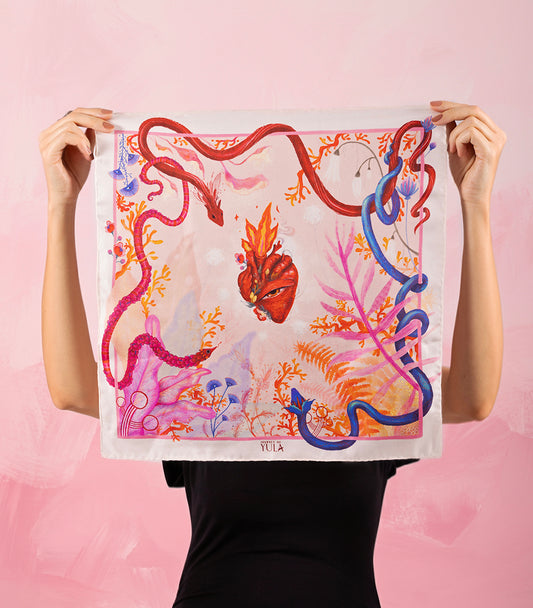 A model holding the full square "Heart Aflame" silk scarf, displaying the complete surrealist illustration of a flaming heart and colorful serpents on a pink background.