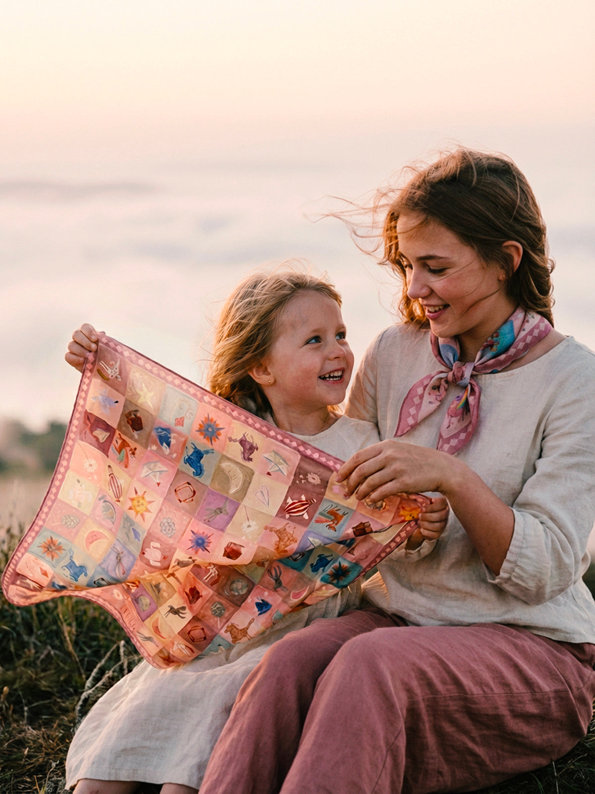 A close-up shot of two young sisters sitting on a grassy hill above a sea of clouds. They are examining the whimsical pastel illustrations on a Journey of Yula scarf. The older girl wears a small, neatly tied 45cm neckerchief. Soft morning light, natural skin textures, and cinematic atmosphere emphasizing a 'pocket of joy' moment.