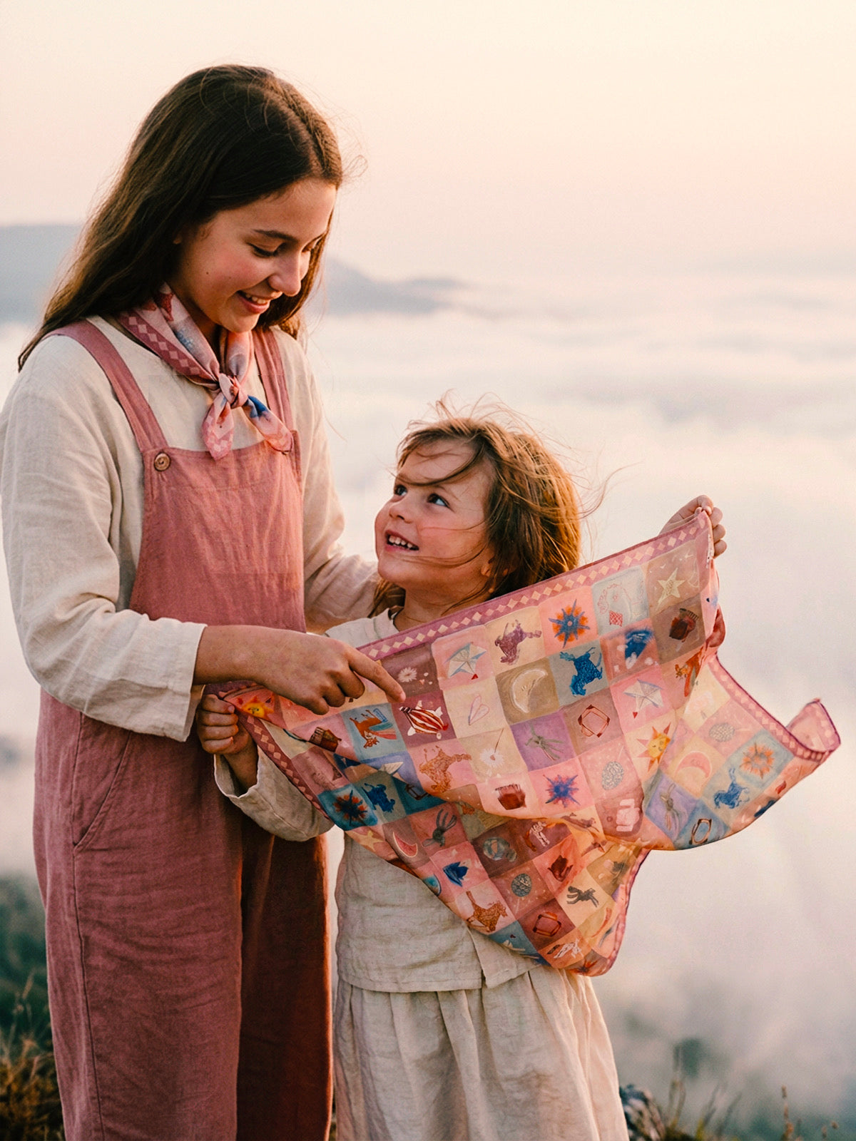 Two sisters with realistic, characteristic features standing on a misty mountain peak at dawn. The younger girl holds a 45cm Journey of Yula silk-cotton scarf spread out like a dream map, while the older sister wears a matching compact neckerchief over pink linen overalls. High-end editorial children's fashion photography