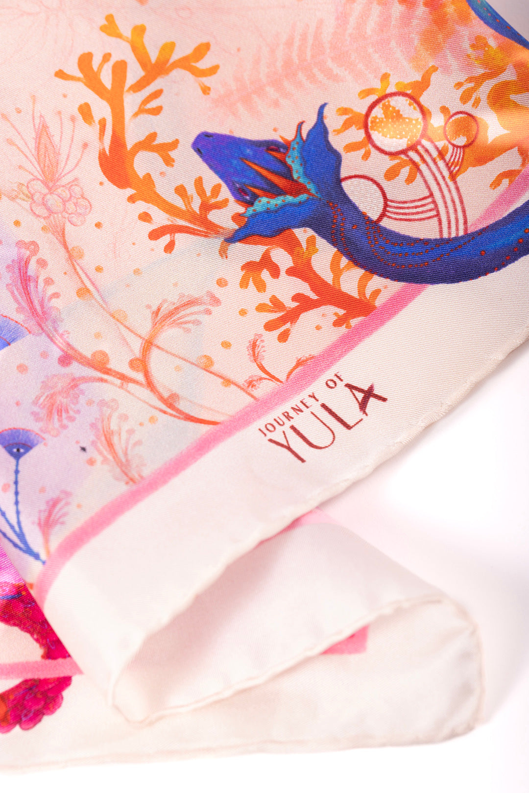 Close-up of the "Journey of Yula" logo printed on the hem of a high-quality silk scarf with hand-rolled edges and detailed botanical illustrations.