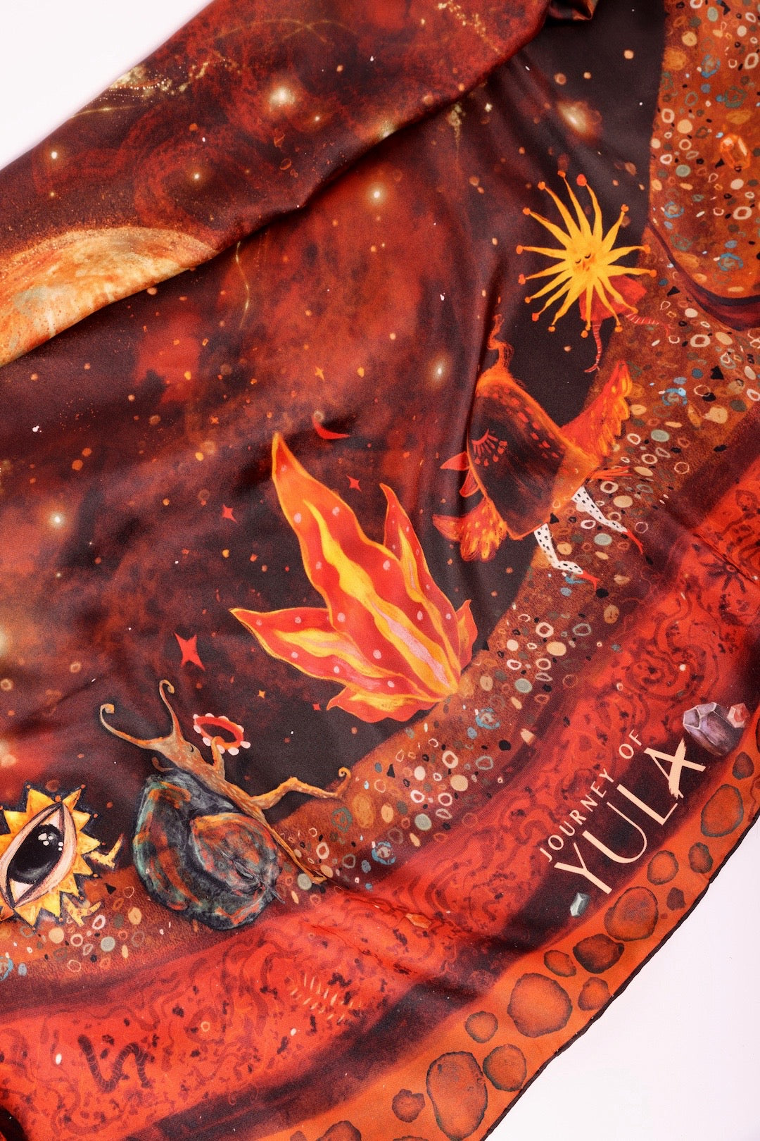 Detailed close-up of a premium 'Journey of Yula' silk scarf corner with vibrant celestial and creature illustrations against a dark cosmic background, including the gold brand logo.