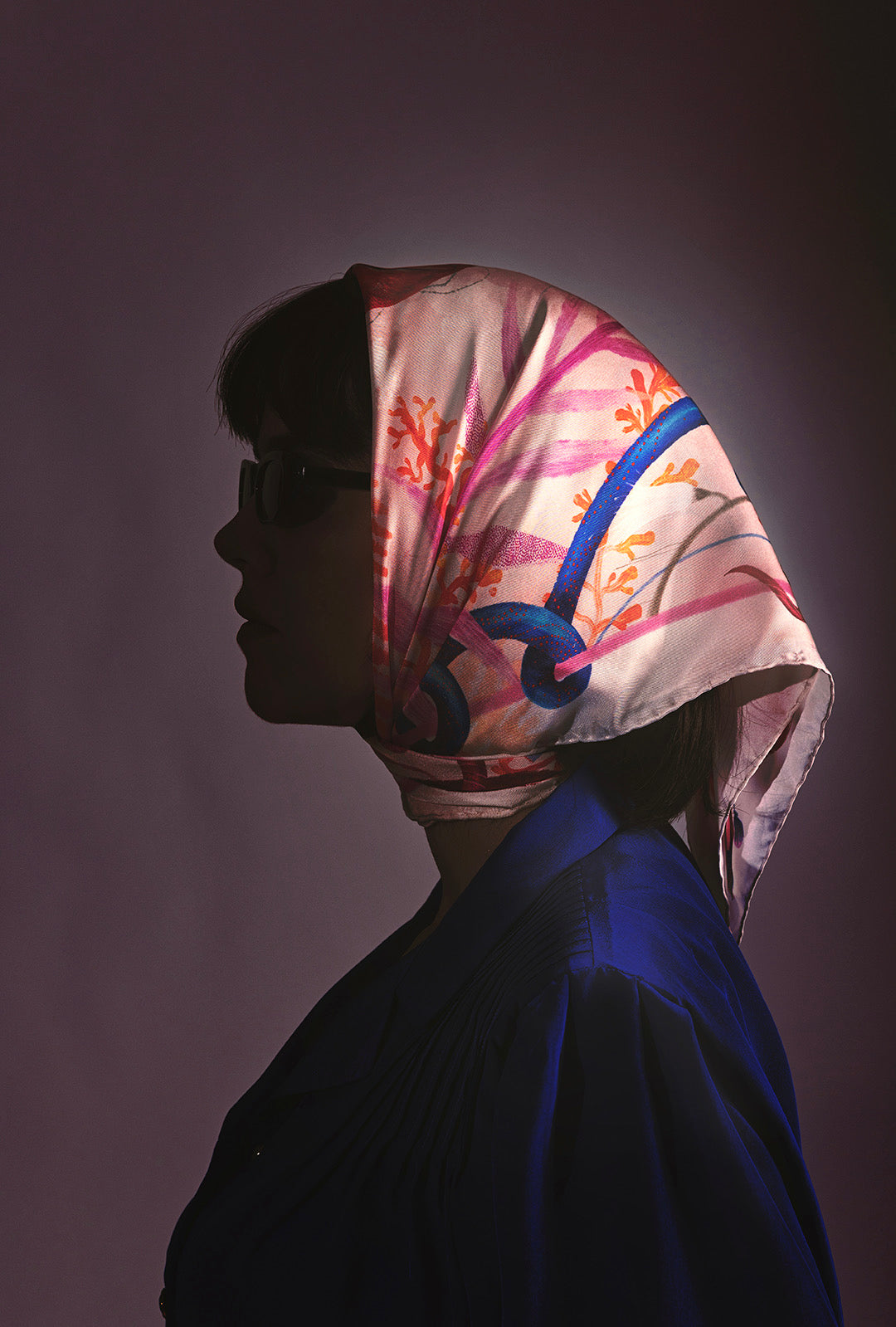 A dramatic profile silhouette of a woman wearing the patterned silk scarf as a headscarf, paired with a dark blue blouse under artistic lighting.