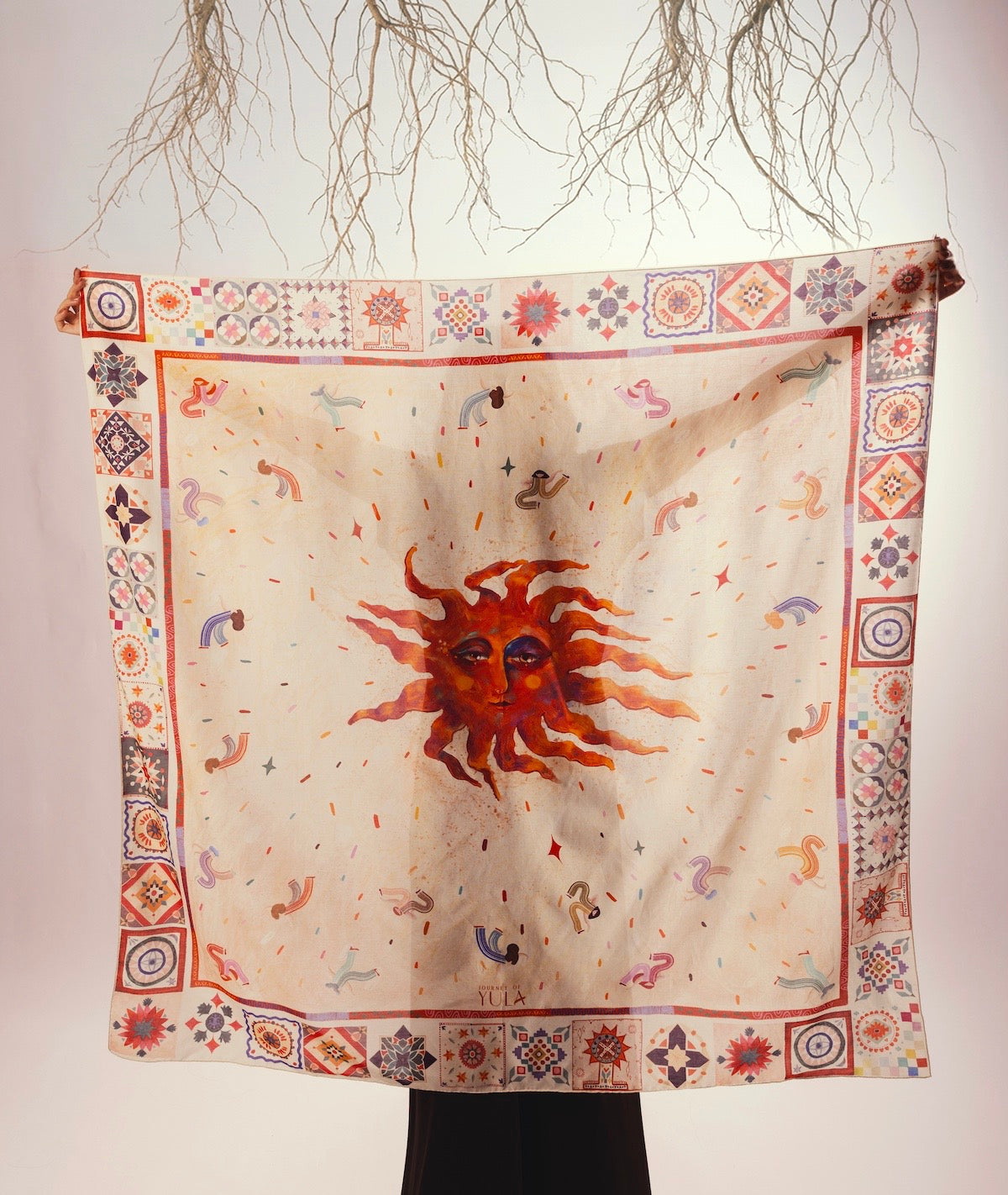 Large square silk twill scarf with a central artistic sun face illustration, ethnic border patterns, and warm cream tones, displayed as wearable art.