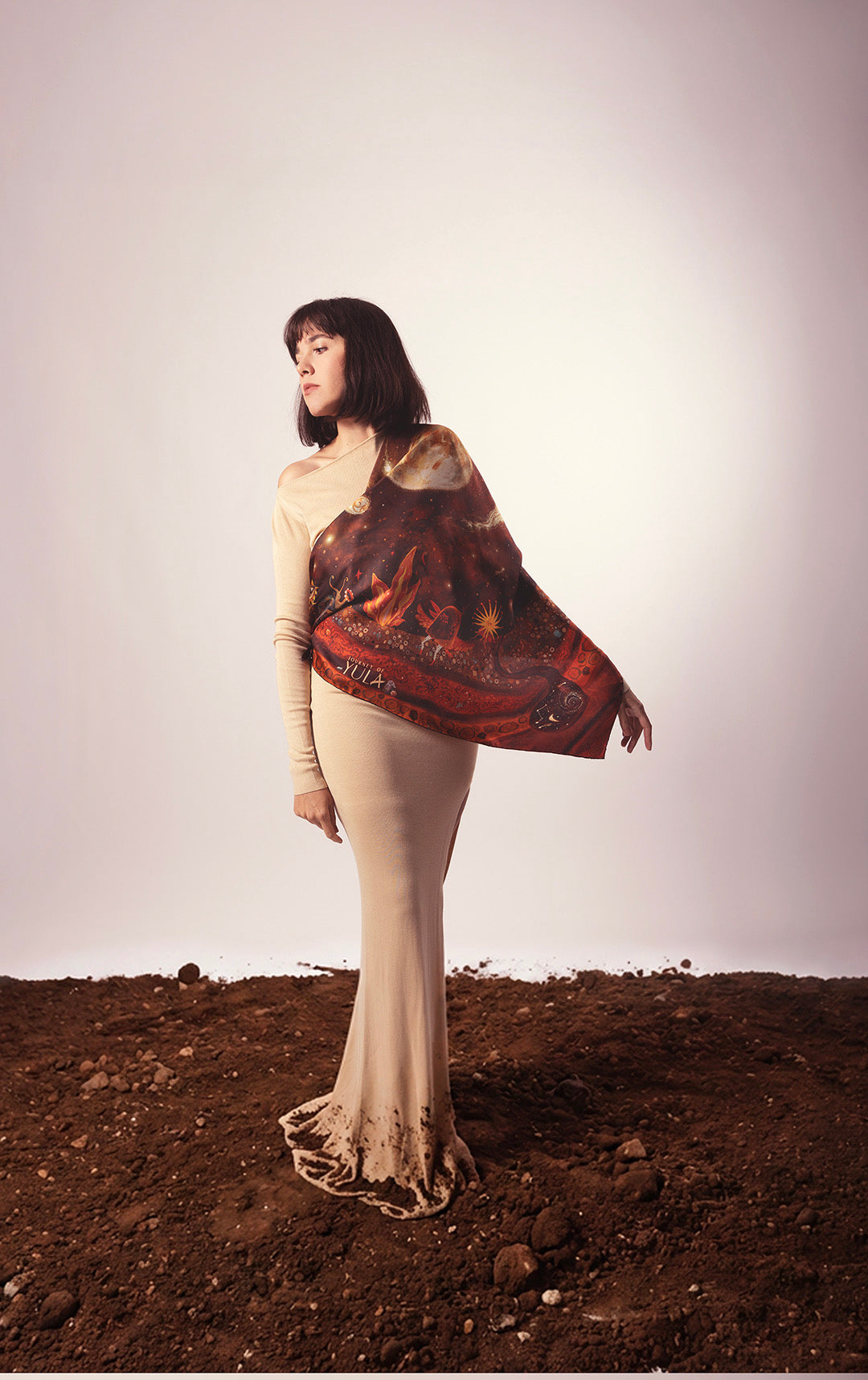 Model wearing a luxury silk scarf with celestial illustrations and a beige dress standing on soil.