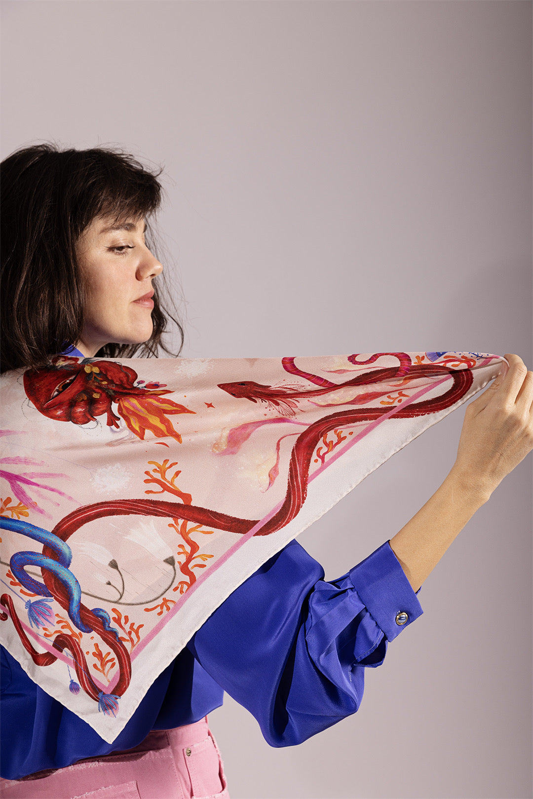 A woman in a blue blouse and pink trousers holding out one corner of the scarf draped over her shoulder to show the drape and silky texture of the fabric.
