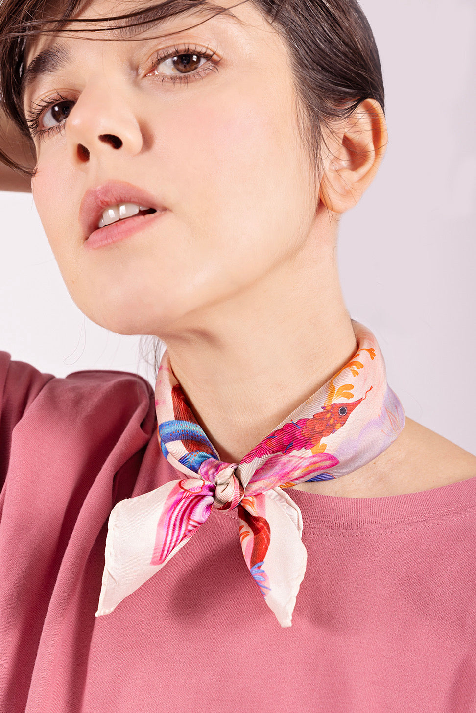 Close-up of a woman wearing a small silk scarf tied closely around the neck, highlighting the vibrant pink serpent and floral motifs.