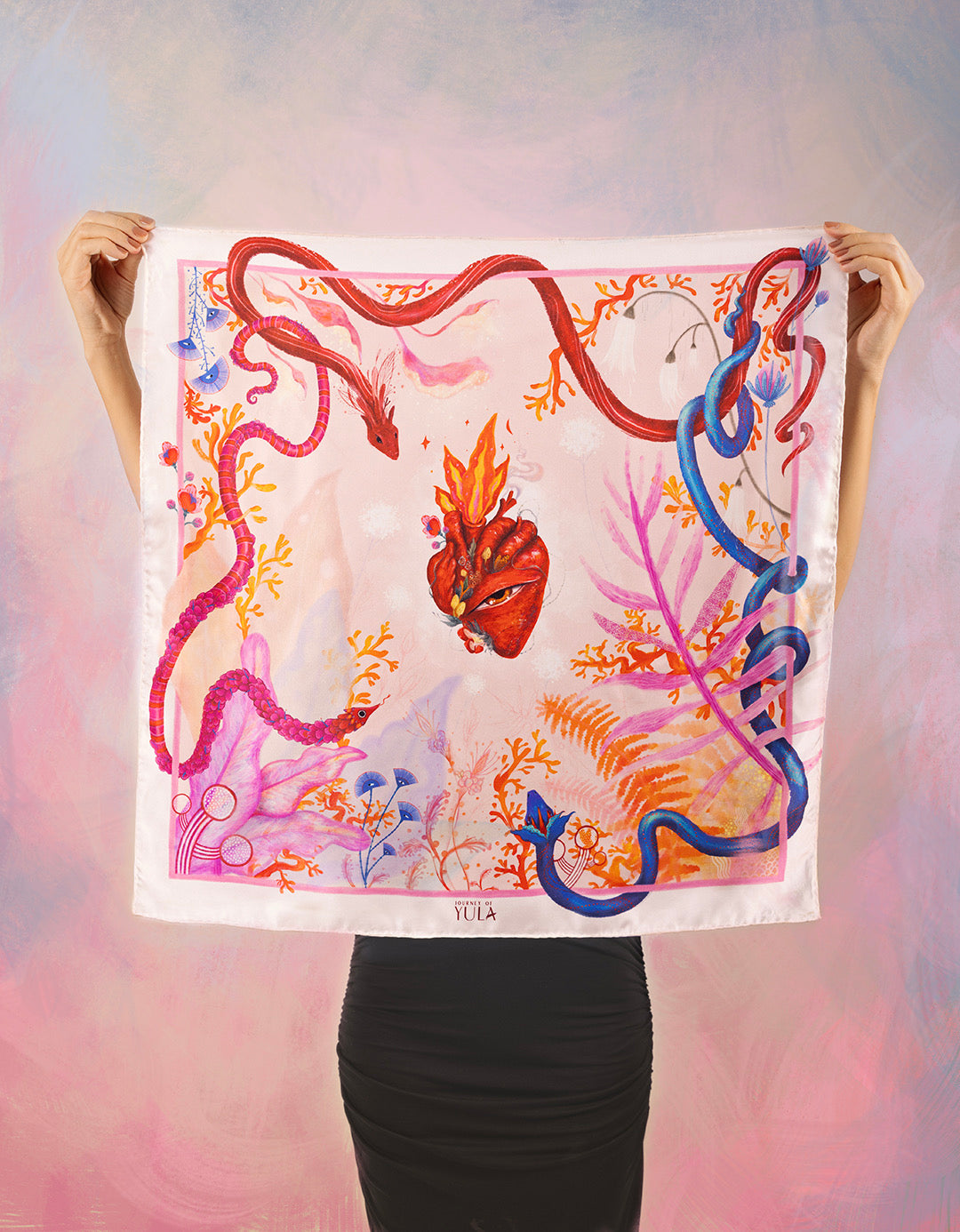 A full view of the square silk scarf held open by two hands, displaying the intricate central heart illustration and the vibrant, symmetrical corner details.