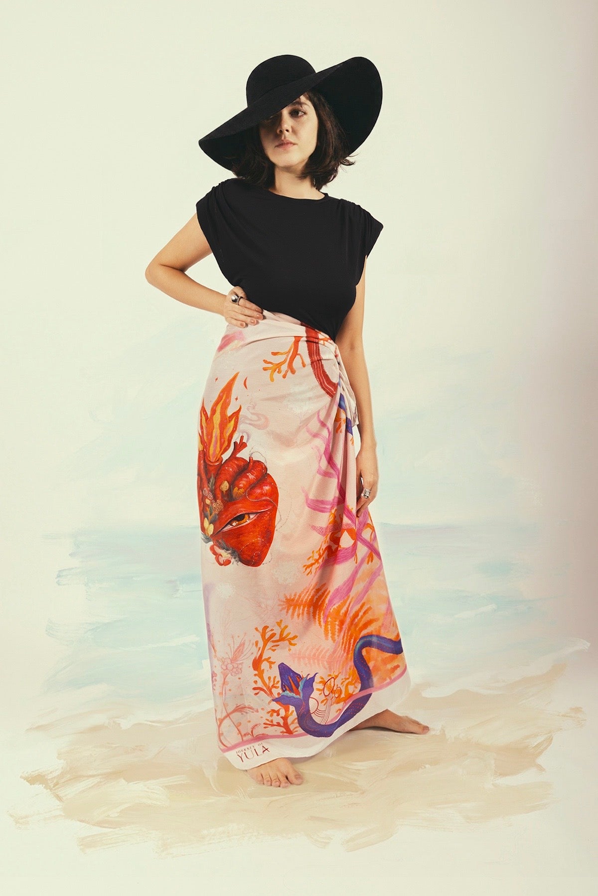 Woman wearing a long pink silk pareo skirt featuring a hand-illustrated flaming heart and snake design, styled with a black hat.