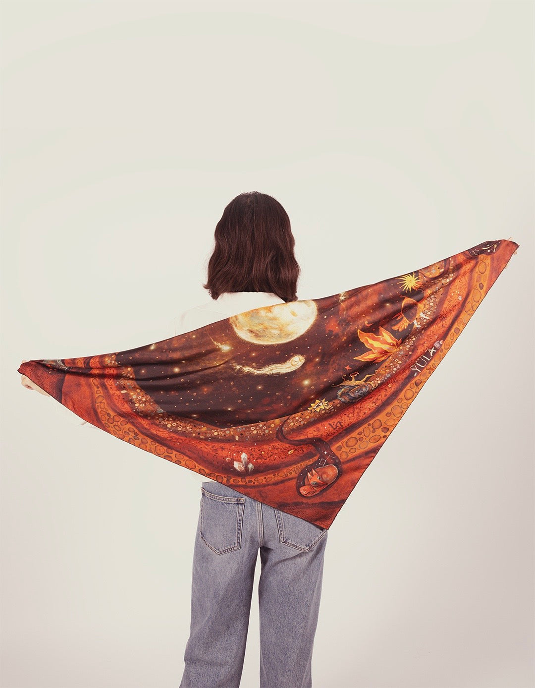 Back view of a model in denim jeans holding up a large illustrated silk scarf with earth tones.