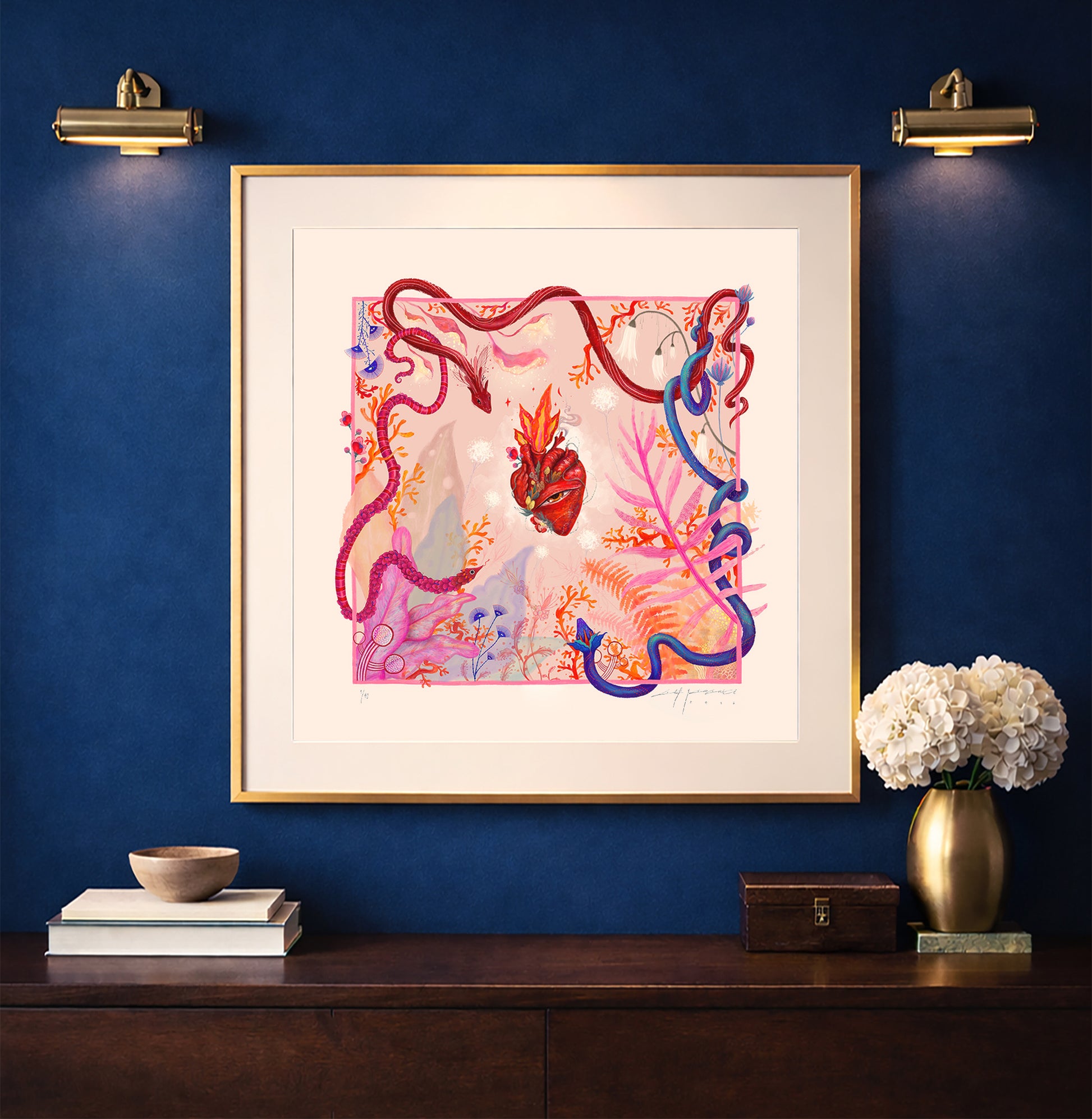 A framed "Heart Aflame" art print displayed on a navy blue wall above a wooden console, illuminated by warm gallery lighting in a modern interior setting.